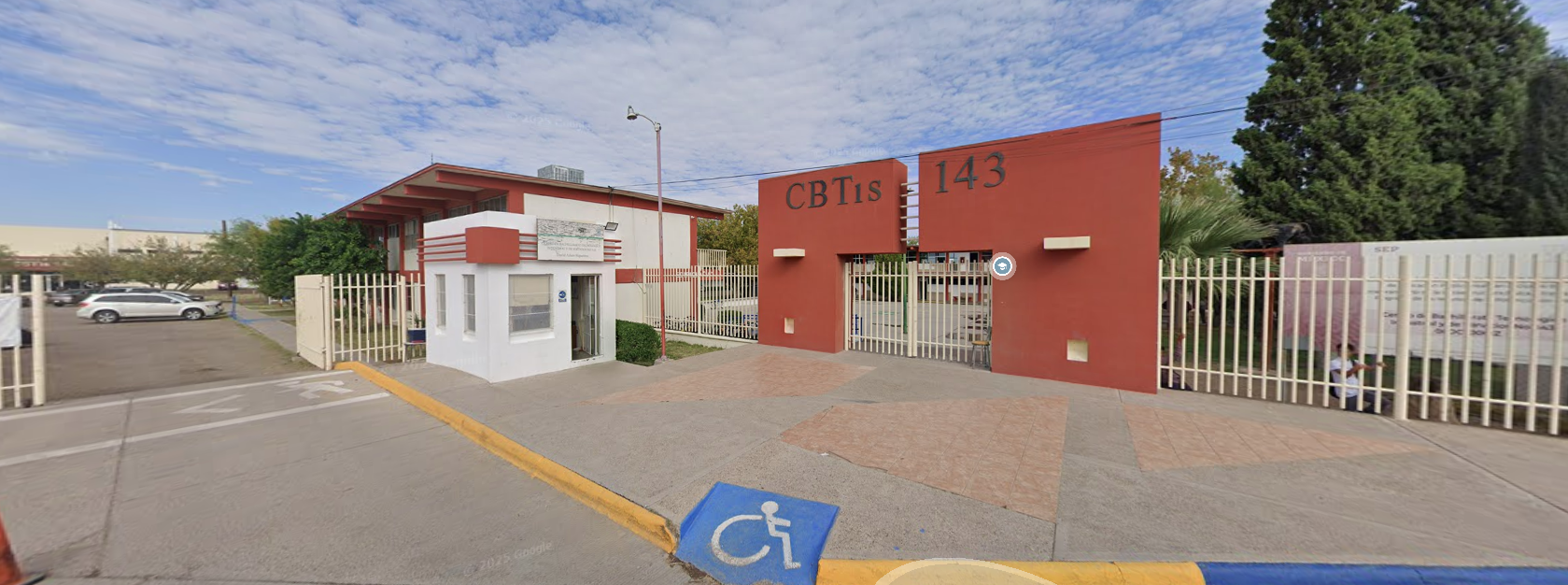 Fotografía en la que se muestra la explanada con una rampa de acceso al plantel. Al fondo de la explanada se encuentra la entrada que está conformada por unos muros de color rojo. Sobre los muros, en la parte superior, hay unas letras grandes negras que dicen Cebetis 143. 